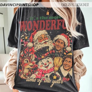 K&ouml;nnte beinhalten: Ein anthrazitfarbenes Kurzarm-T-Shirt mit einem Grafikdruck und dem Text 'it's the most WONDERFUL time of the year'. Das Design zeigt Illustrationen vom Weihnachtsmann, einem Rentier, einem Schneemann und Buddy dem Elf.