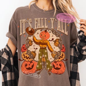 May include: Brown t-shirt with the text "IT'S FALL Y'ALL" and a pumpkin-headed cowboy graphic. The graphic includes pumpkins, bats, and a rope design. The cowboy is wearing a yellow shirt, fringed vest, and green pants with stars.