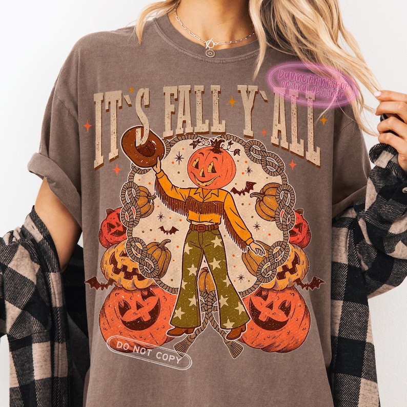 May include: Brown t-shirt with the text "IT'S FALL Y'ALL" and a pumpkin-headed cowboy graphic. The graphic includes pumpkins, bats, and a rope design. The cowboy is wearing a yellow shirt, fringed vest, and green pants with stars.