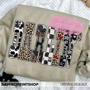 May include: A beige sweatshirt with the word "DARLIN" in a bold, multi-patterned design. The letters feature cow print, leopard print, stars, and checkered patterns. The text "DaVinciPrintShop Original Designer" is also visible.