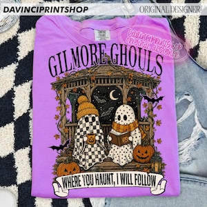 Puede incluir: Una camiseta de color morado claro con un gr&aacute;fico de dos fantasmas en un cenador. Un fantasma lleva un gorro y sostiene una taza, mientras el otro lee un libro. El dise&ntilde;o incluye calabazas, murci&eacute;lagos y el texto "GILMORE GHOULS" y "WHERE YOU HAUNT, I WILL FOLLOW."