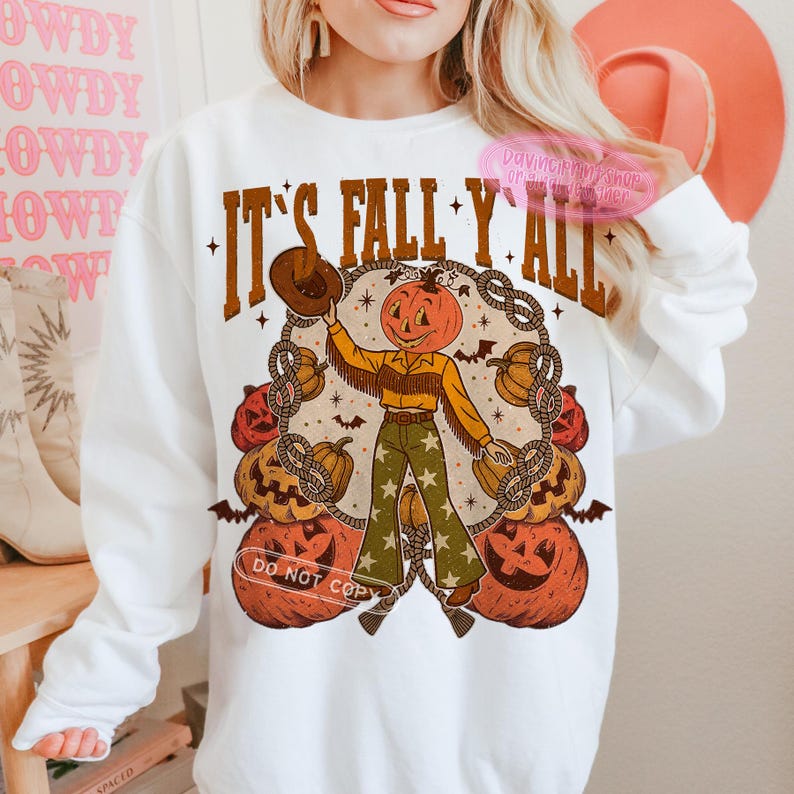 May include: White sweatshirt with a fall-themed graphic. The design features a pumpkin-headed cowboy holding a hat, surrounded by pumpkins and rope, with the text "IT'S FALL Y'ALL". The design is in shades of brown, orange, and green.