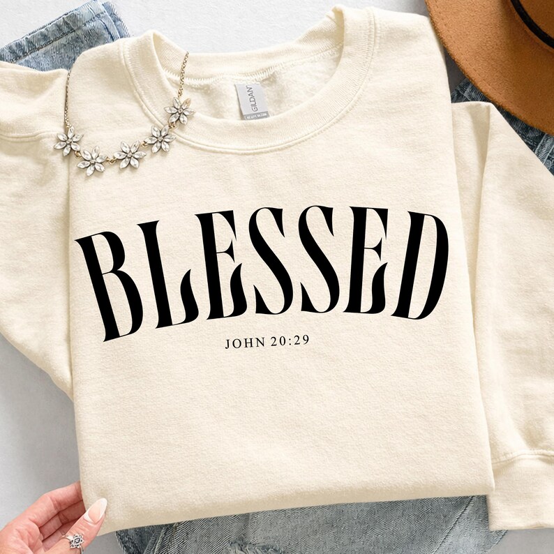 K&ouml;nnte beinhalten: Cremefarbenes Sweatshirt mit dem Wort "BLESSED" in gro&szlig;en, schwarzen, fetten Buchstaben. Darunter steht der Text "JOHN 20:29". Eine zarte Blumenkette und ein brauner Hut sind ebenfalls zu sehen.