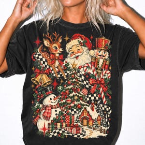 May include: A black short-sleeve t-shirt featuring a colorful Christmas graphic. The design includes Santa Claus, a reindeer, a nutcracker, a snowman, a decorated tree, wrapped gifts, a small house, bells, and candy canes with a checkered ribbon.
