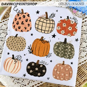 May include: A white t-shirt featuring a graphic print of nine pumpkins in various patterns and colors, including leopard print, plaid, floral, polka dot, and heart motifs, interspersed with small black stars and vine-like flourishes.