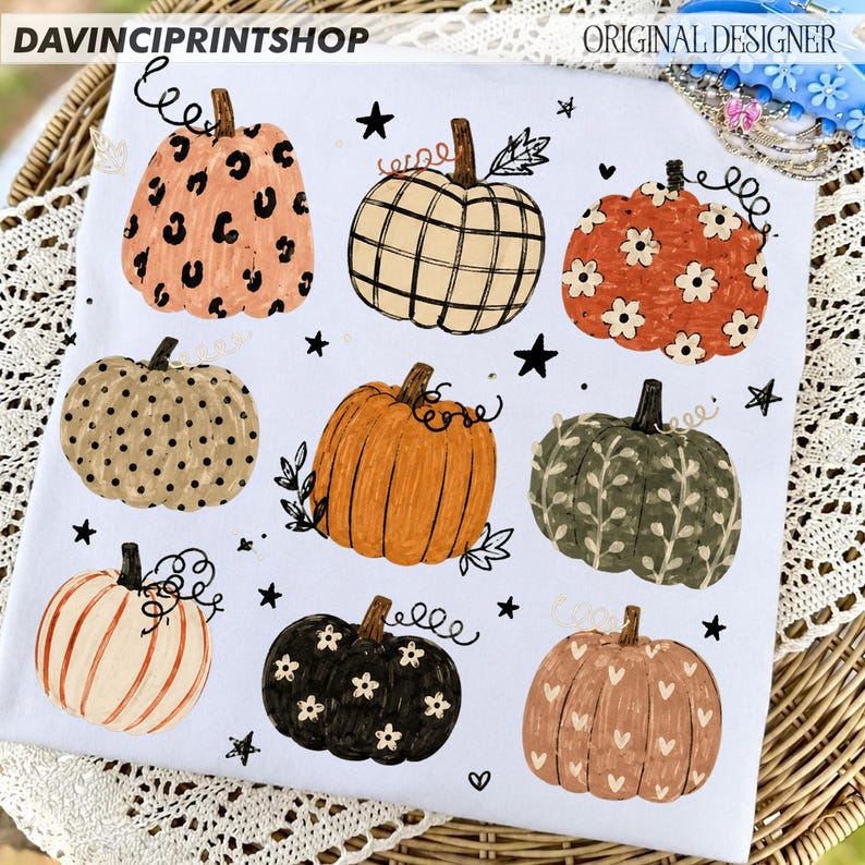 May include: A white t-shirt featuring a graphic print of nine pumpkins in various patterns and colors, including leopard print, plaid, floral, polka dot, and heart motifs, interspersed with small black stars and vine-like flourishes.
