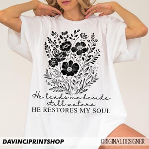 May include: White t-shirt with a black floral design and the text "He leads me beside still waters HE RESTORES MY SOUL".