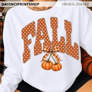 May include: A white long-sleeve sweatshirt featuring the word FALL in large, orange, white-polka-dotted letters with a leopard print outline. Below the text, two orange pumpkins are tied together with a brown and white plaid ribbon bow.