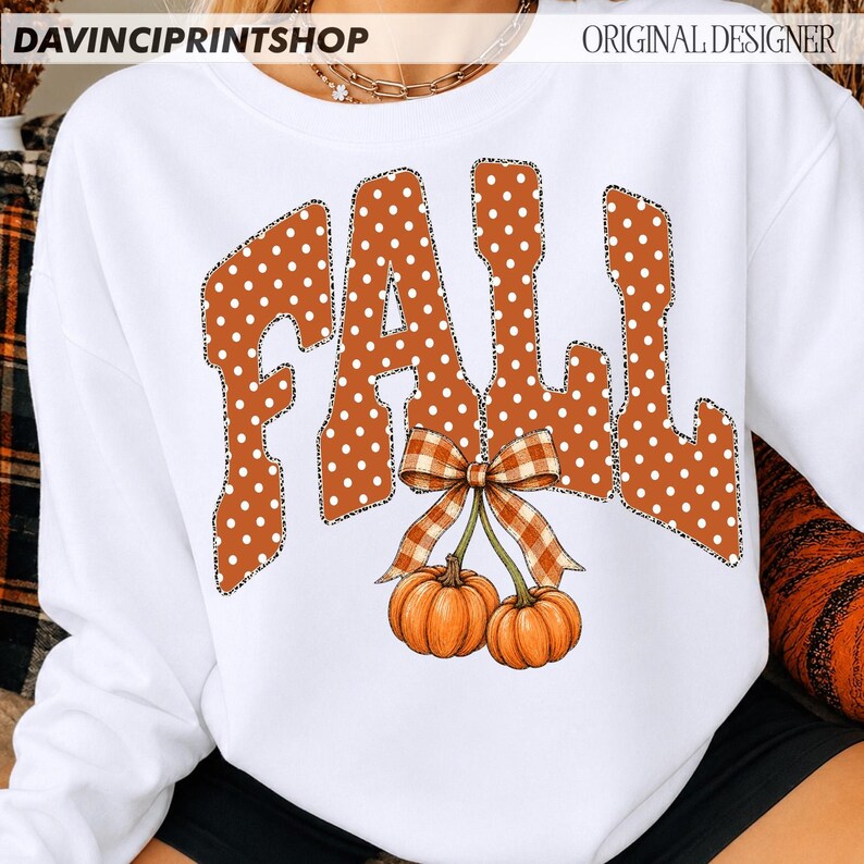May include: A white long-sleeve sweatshirt featuring the word FALL in large, orange, white-polka-dotted letters with a leopard print outline. Below the text, two orange pumpkins are tied together with a brown and white plaid ribbon bow.