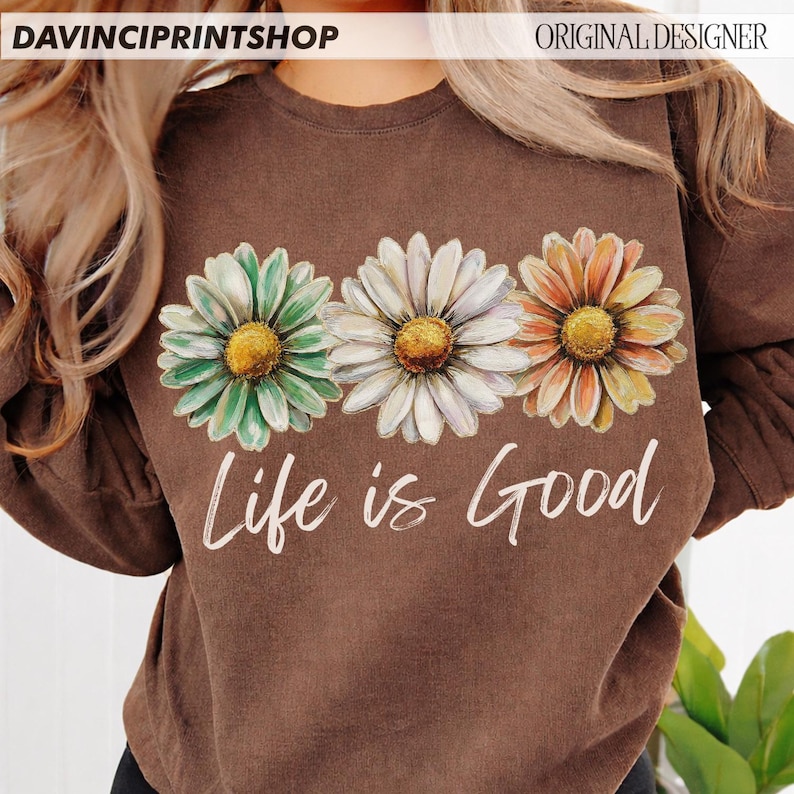 May include: A brown long-sleeved top featuring three daisy flowers in green, white, and orange. The words "Life is Good" are printed in white script below the flowers. The top has a crew neck.