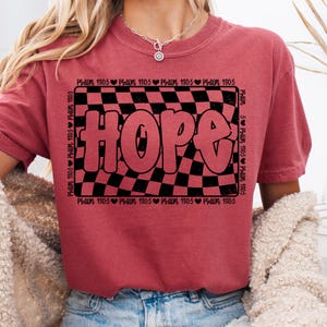Op de afbeelding: Rood T-shirt met een zwart-wit geruit patroon en het woord "HOPE" in grote, roze letters. Het geruite patroon is omgeven door een zwarte rand met de tekst "Psalm 130:5" die rond de rand wordt herhaald.