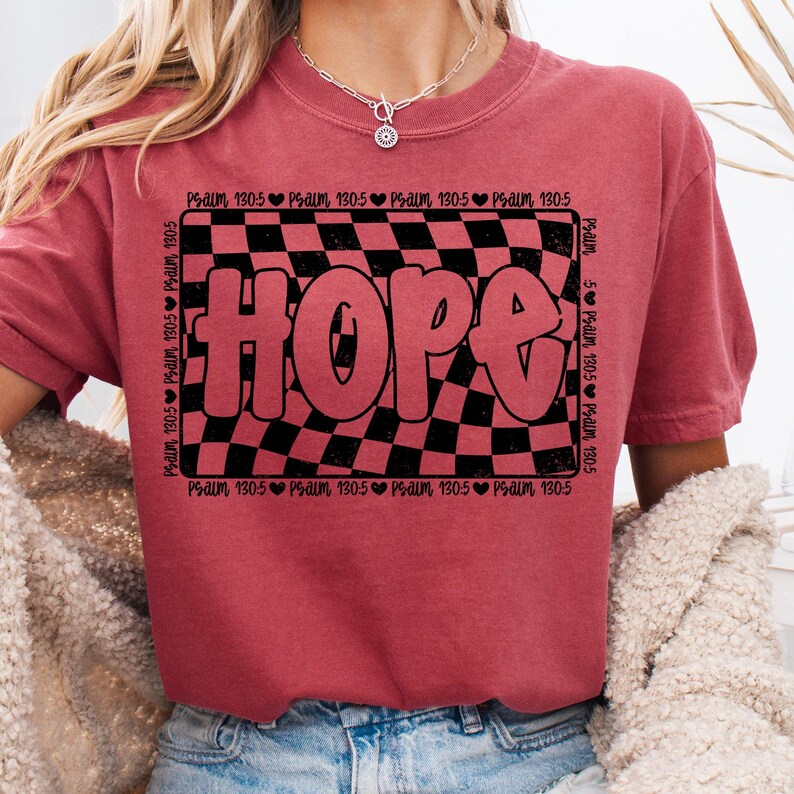 Op de afbeelding: Rood T-shirt met een zwart-wit geruit patroon en het woord "HOPE" in grote, roze letters. Het geruite patroon is omgeven door een zwarte rand met de tekst "Psalm 130:5" die rond de rand wordt herhaald.