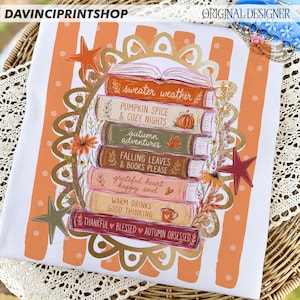 K&ouml;nnte beinhalten: Ein wei&szlig;es T-Shirt mit einem Stapel aus sechs B&uuml;chern mit orangefarbenen, gr&uuml;nen, braunen und rosafarbenen Buchr&uuml;cken. Die Buchtitel lauten: sweater weather, PUMPKIN SPICE & COZY NIGHTS, autumn adventures, FALLING LEAVES & BOOKS PLEASE, grateful heart happy soul, WARM DRINKS GOOD THINKING und THANKFUL BLESSED AUTUMN OBSESSED.