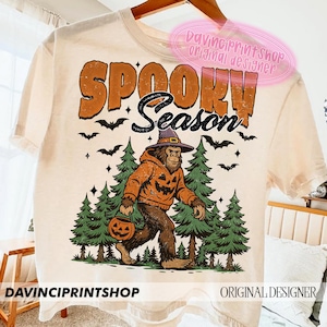 May include: Cream-colored t-shirt with the words "SPOOKY SEASON" in orange and black. A Bigfoot-like creature wearing a pumpkin hoodie and witch hat walks through a forest, carrying a jack-o'-lantern. Bats and stars are scattered around the design.