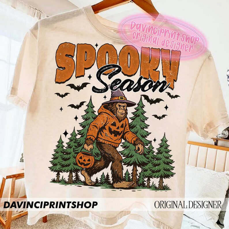 May include: Cream-colored t-shirt with the words "SPOOKY SEASON" in orange and black. A Bigfoot-like creature wearing a pumpkin hoodie and witch hat walks through a forest, carrying a jack-o'-lantern. Bats and stars are scattered around the design.