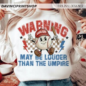 K&ouml;nnte beinhalten: Wei&szlig;es Sweatshirt mit Baseball-Motiv. Das Design zeigt eine Cartoon-Baseballfigur mit roter Kappe und Schl&auml;ger, mit dem Text "WARNING GAME DAY" und "MAY BE LOUDER THAN THE UMPIRE".