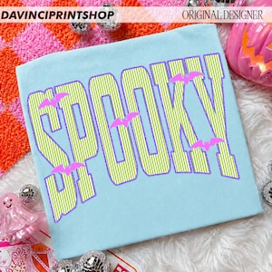 May include: A light blue t-shirt featuring the word SPOOKY in large, lime green and white vertical striped letters with a purple outline. Small, solid pink bat silhouettes are positioned between and around the letters of the text.