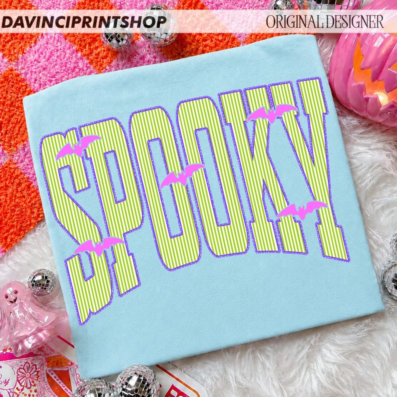 May include: A light blue t-shirt featuring the word SPOOKY in large, lime green and white vertical striped letters with a purple outline. Small, solid pink bat silhouettes are positioned between and around the letters of the text.