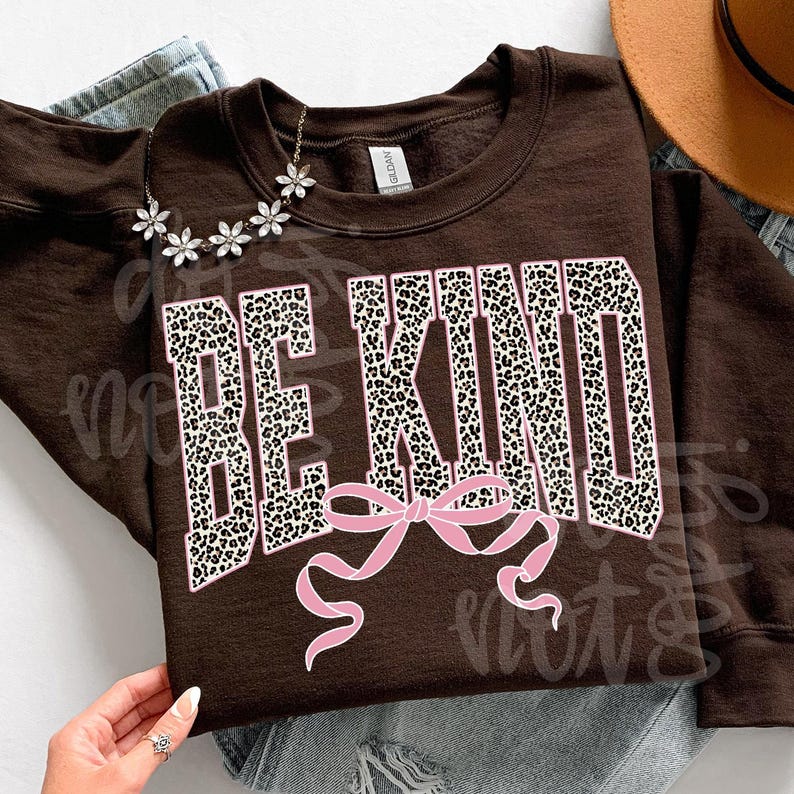 Op de afbeelding: Een bruine sweatshirt met de tekst "BE KIND" in een wit en luipaardprint ontwerp, met een roze strik. Een bloemenketting ligt op de sweatshirt. De sweatshirt ligt op een blauwe jeans.