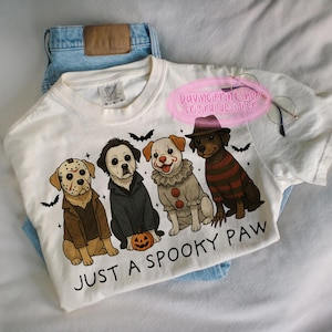 May include: White t-shirt with a Halloween-themed graphic featuring four dogs dressed as horror movie characters. The phrase "JUST A SPOOKY PAW" is printed below the dogs. The shirt is laid on top of blue jeans.