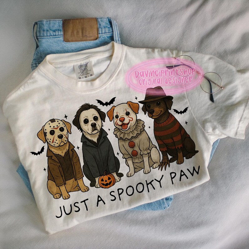 May include: White t-shirt with a Halloween-themed graphic featuring four dogs dressed as horror movie characters. The phrase "JUST A SPOOKY PAW" is printed below the dogs. The shirt is laid on top of blue jeans.