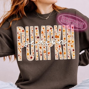 May include: A dark gray crewneck sweatshirt featuring the word PUMPKIN in large block letters filled with a floral pattern of orange, yellow, and brown flowers and small pumpkins, with the word season written in white cursive script across the center.