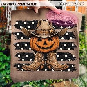 K&ouml;nnte beinhalten: Ein braunes T-Shirt mit einem Grafikdruck eines K&uuml;rbisses, der einen Cowboyhut und Cowboystiefel tr&auml;gt. Das Design ist vor einem Hintergrund aus schwarzen horizontalen Pinselstrichen mit wei&szlig;en Punkten platziert. Der Text DAVINCIPRINTSHOP ist sichtbar.