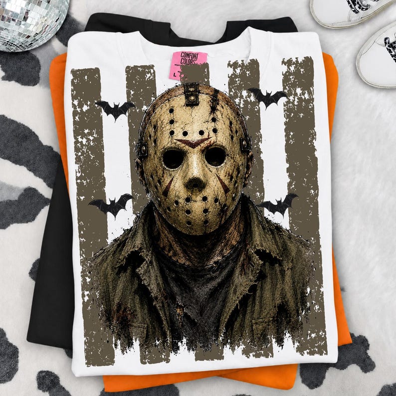 May include: A white t-shirt featuring a distressed graphic of a hockey mask with dark eye holes and ventilation dots. The design includes vertical textured stripes and black bat silhouettes. A pink Comfort Colors brand tag is visible at the neck.