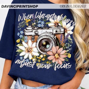 May include: Navy blue t-shirt featuring a vintage camera design surrounded by colorful flowers. The text "When life gets blurry adjust your focus" is written in white script. The shirt has the text "DAVINCIPRINTSHOP" and "ORIGINAL DESIGNER" at the top.