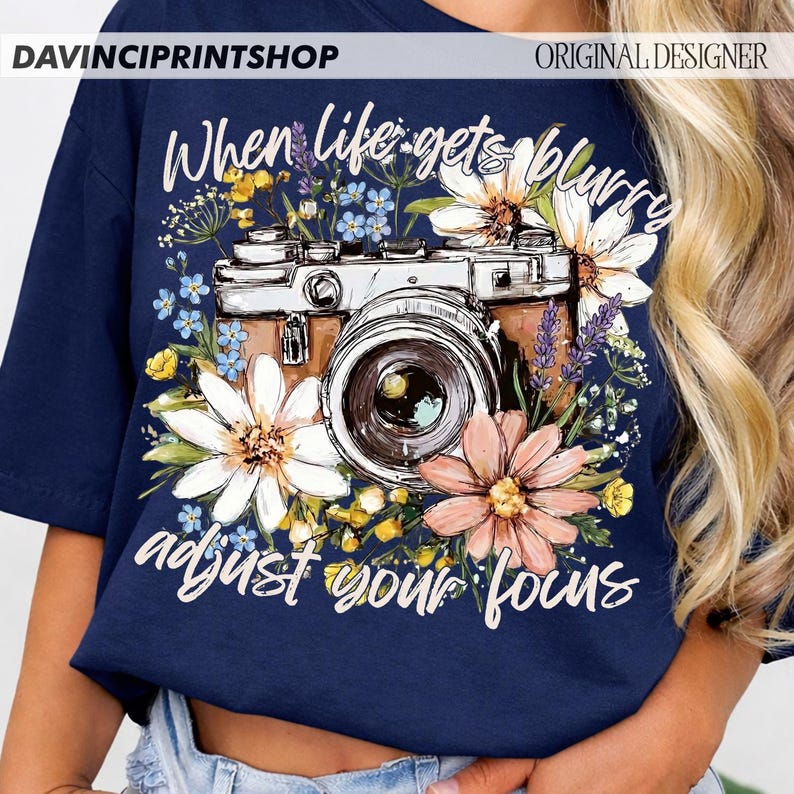 May include: Navy blue t-shirt featuring a vintage camera design surrounded by colorful flowers. The text "When life gets blurry adjust your focus" is written in white script. The shirt has the text "DAVINCIPRINTSHOP" and "ORIGINAL DESIGNER" at the top.