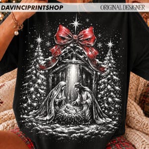 May include: A black t-shirt featuring a graphic print of a nativity scene with a large red bow at the top. The design includes two figures standing over a manger, flanked by two decorated Christmas trees, all set against a starry night background.