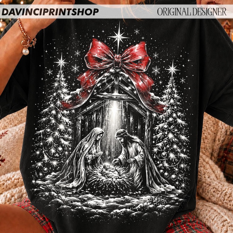 May include: A black t-shirt featuring a graphic print of a nativity scene with a large red bow at the top. The design includes two figures standing over a manger, flanked by two decorated Christmas trees, all set against a starry night background.