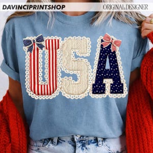 May include: Light blue t-shirt with the word "USA" in a patriotic design. The "U" is red and white striped, the "S" is a cream cable knit, and the "A" is navy blue with white stars. Each letter is outlined with a white lace trim and topped with a bow.