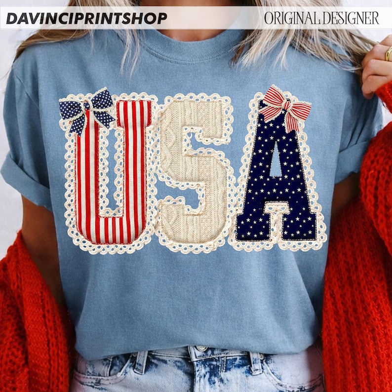 May include: Light blue t-shirt with the word "USA" in a patriotic design. The "U" is red and white striped, the "S" is a cream cable knit, and the "A" is navy blue with white stars. Each letter is outlined with a white lace trim and topped with a bow.