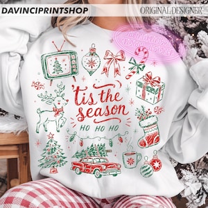 May include: A white sweatshirt featuring a red and green Christmas-themed graphic print with the text 'tis the season' and 'HO HO HO'. The design includes a vintage television, reindeer, gift box, stocking, car, and various holiday ornaments.