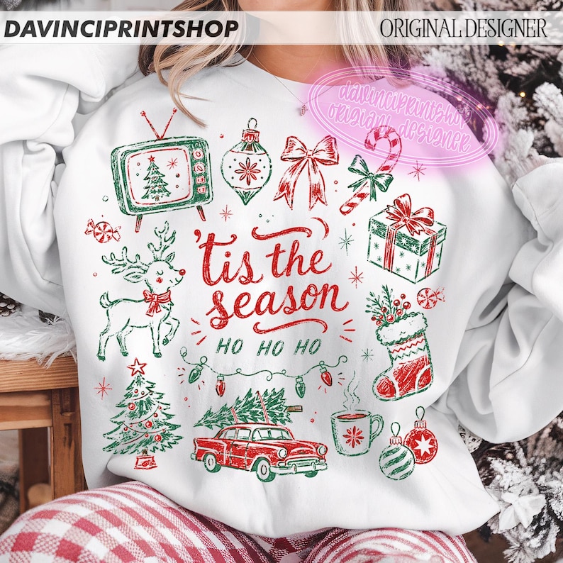 May include: A white sweatshirt featuring a red and green Christmas-themed graphic print with the text 'tis the season' and 'HO HO HO'. The design includes a vintage television, reindeer, gift box, stocking, car, and various holiday ornaments.