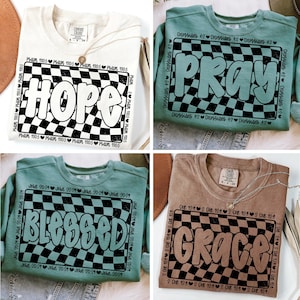 Op de afbeelding: Vier verschillende sweatshirts met een geruit patroon en een woord in een vetgedrukt, bubbelvormig lettertype. De woorden zijn "Hope", "Pray", "Blessed" en "Grace". Elk sweatshirt heeft een andere kleur en een ander bijbelvers dat rond het geruite patroon is gedrukt.