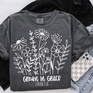 Pode incluir: Uma camiseta cinza-chumbo com um gr&aacute;fico branco de flores e o texto "Grown in Grace 2 Peter 3:18".