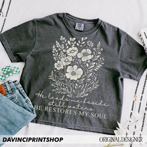 May include: A charcoal gray t-shirt with a floral design and the text "He leads me beside still waters HE RESTORES MY SOUL".
