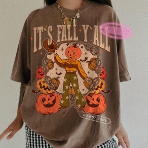 May include: Brown t-shirt with the text "IT'S FALL Y'ALL" in a vintage style. The graphic features a pumpkin-headed cowboy surrounded by pumpkins and rope. The cowboy is wearing a hat, fringe jacket, and star-patterned pants.