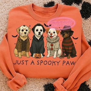 May include: Orange sweatshirt with a graphic of four dogs dressed as horror movie characters. The dogs are wearing costumes of Jason Voorhees, Michael Myers, Pennywise, and Freddy Krueger. The text "JUST A SPOOKY PAW" is below the dogs.