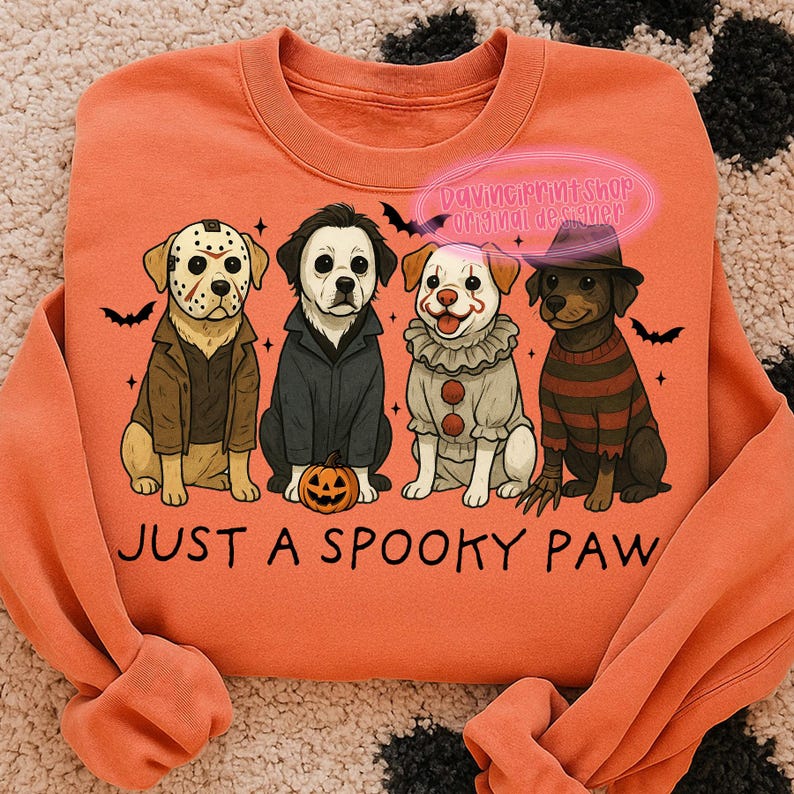 May include: Orange sweatshirt with a graphic of four dogs dressed as horror movie characters. The dogs are wearing costumes of Jason Voorhees, Michael Myers, Pennywise, and Freddy Krueger. The text "JUST A SPOOKY PAW" is below the dogs.