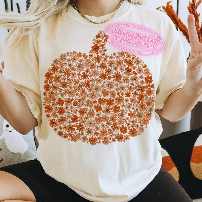 May include: Cream-colored t-shirt featuring a pumpkin design made of orange and peach-colored flowers. The shirt has short sleeves and a crew neck. The text "DaVinciPrintShop original designer" is on the shirt.
