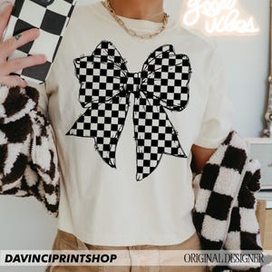 May include: Cream-colored t-shirt featuring a large black and white checkered bow graphic. The shirt has a relaxed fit. The text "DAVINCIPRINTSHOP" is at the bottom of the shirt. The words "ORIGINAL DESIGNER" are also visible.