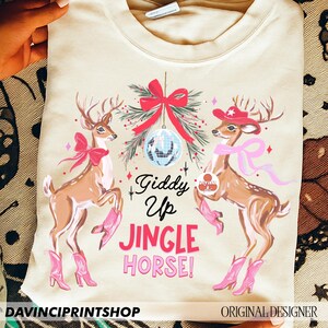 May include: A cream-coloured sweatshirt with a festive design. Two deer wearing cowboy boots and hats are featured, along with a disco ball and the text "Giddy Up Jingle Horse!" A red bow and sprigs of greenery complete the holiday theme.