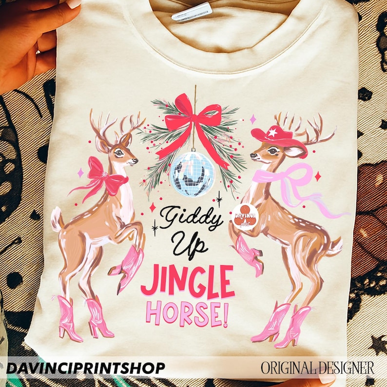 May include: A cream-coloured sweatshirt with a festive design. Two deer wearing cowboy boots and hats are featured, along with a disco ball and the text "Giddy Up Jingle Horse!" A red bow and sprigs of greenery complete the holiday theme.