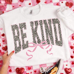 Op de afbeelding: Witte sweatshirt met de tekst "BE KIND" in een luipaardprint met een roze rand. Een roze strik is onder de tekst geplaatst. De sweatshirt staat op een roze en witte geruite achtergrond met hartmotieven.