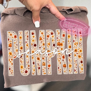 May include: A brown crewneck sweatshirt featuring the word PUMPKIN in large block letters filled with a floral and pumpkin pattern, outlined in white faux pearls. The word season is written in white cursive script across the center.