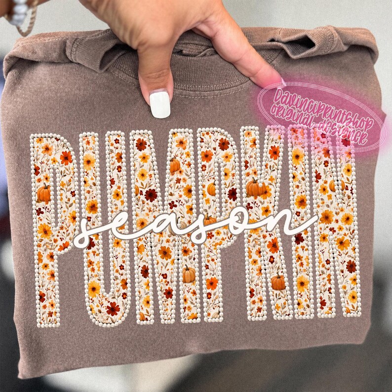 May include: A brown crewneck sweatshirt featuring the word PUMPKIN in large block letters filled with a floral and pumpkin pattern, outlined in white faux pearls. The word season is written in white cursive script across the center.
