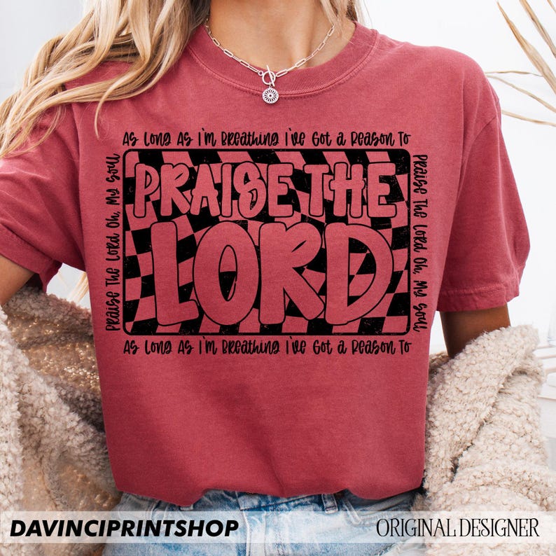 May include: Red t-shirt with a black and white checkered design and the text "Praise The Lord" in a bold, black font. The text "As Long As I'm Breathing I've Got a Reason To" is written in black above and below the main text.